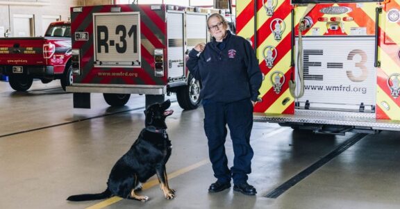 firefighter Charlie standing with fire pup sitting in station with trucks rigs