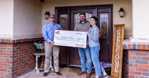 military couple standing outside new home holding big check with Homes for Heroes affiliate showing savings