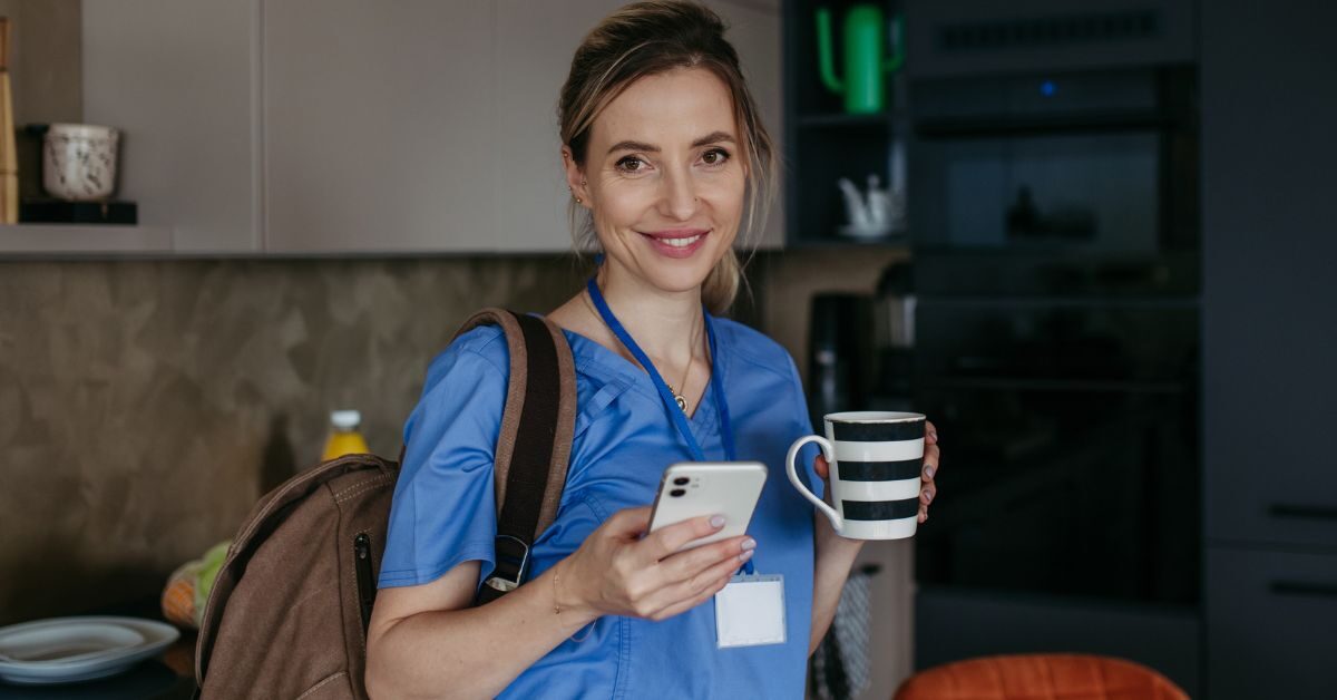 nurse at home in kitchen wearing scrubs holding coffee cell phone carrying backpack smiling